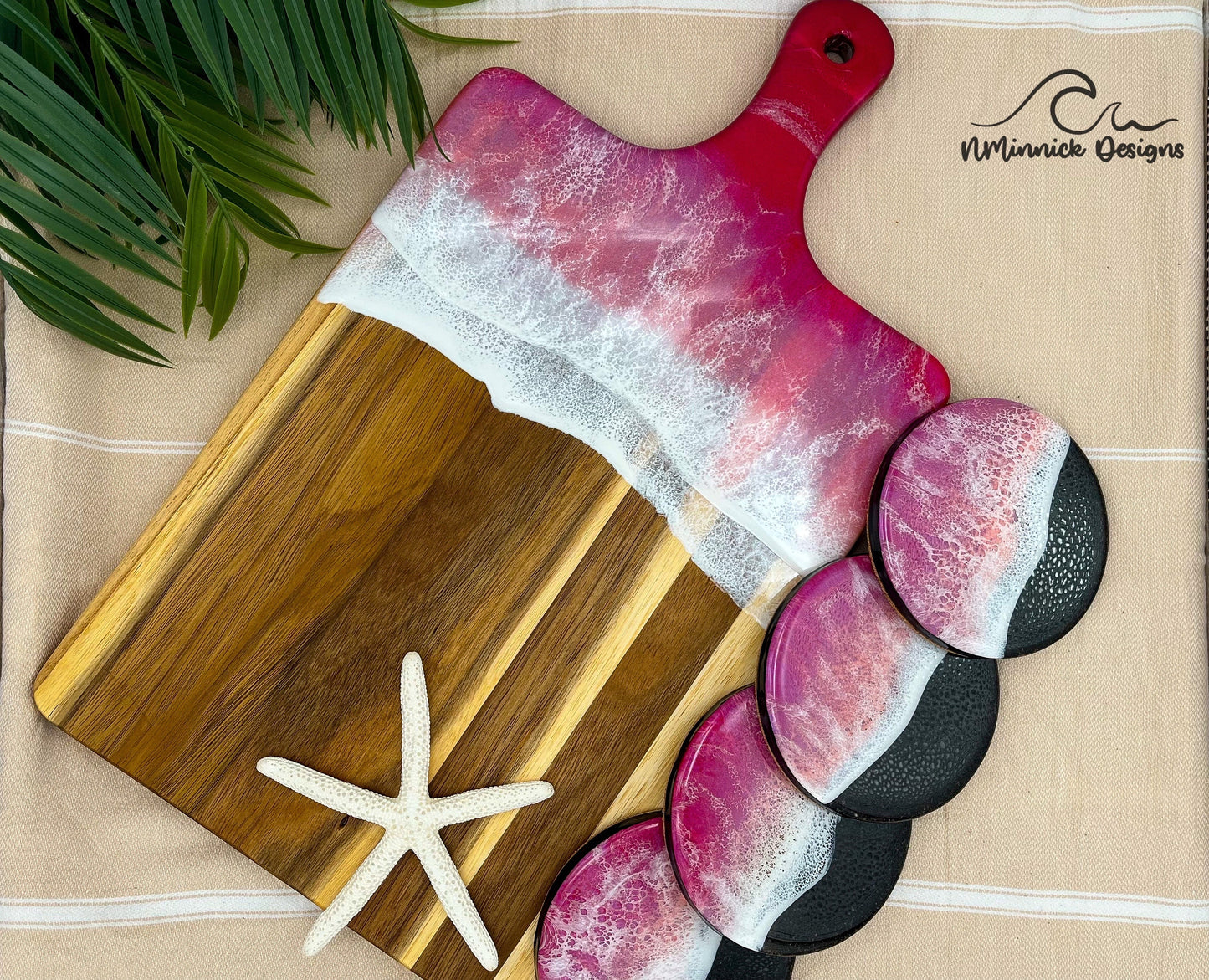 Acacia Wood Serving Board with handle.  Top portion covered in red and pink ocean resin art resembling waves of the ocean.  Matching set of 4 ocean resin drink coasters with real black sand and epoxy resin ocean and cork backings.  Laying on a tan beach towel with a natural starfish (not included).