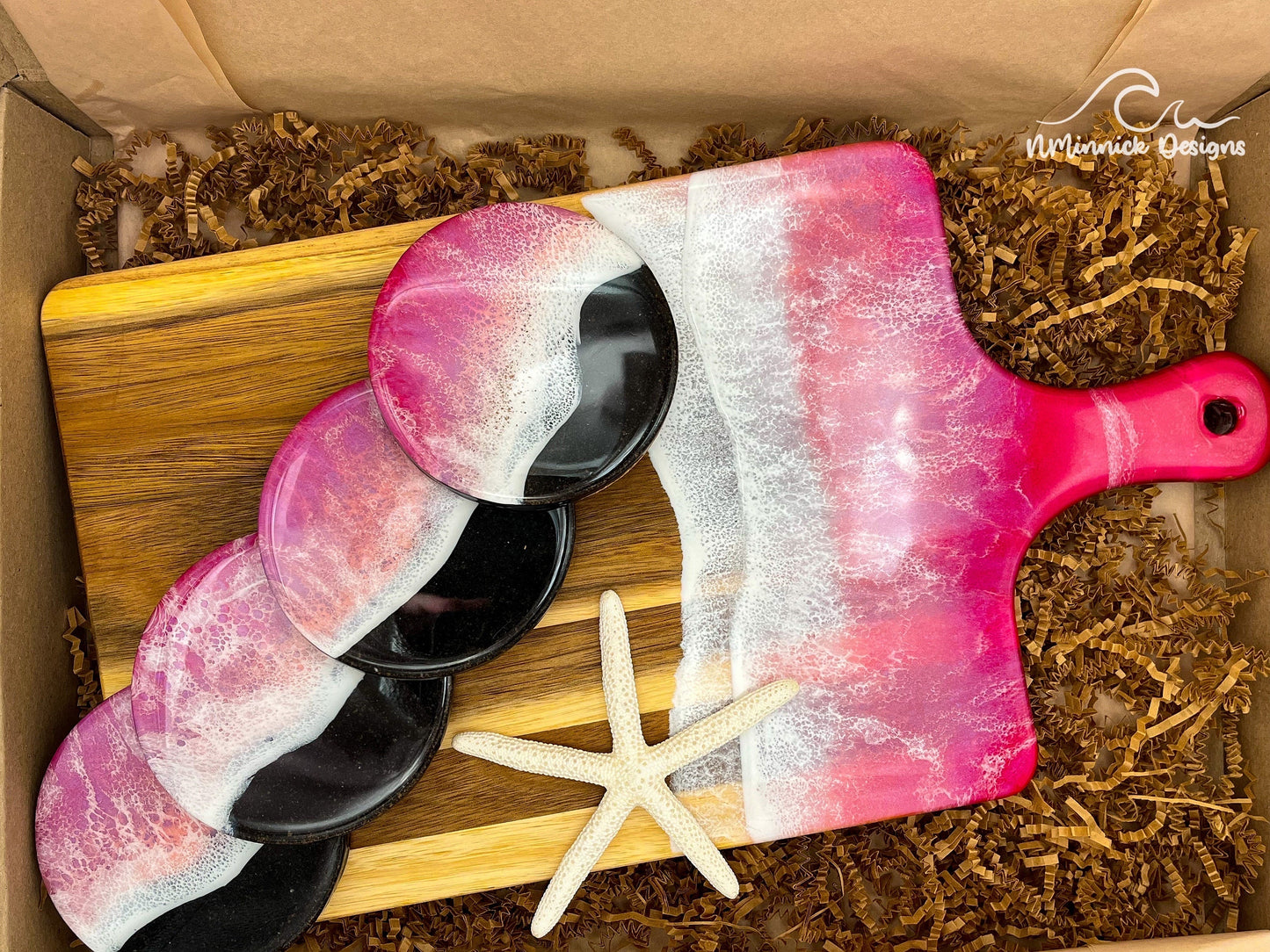 Acacia Wood Serving Board with handle. Top portion covered in red and pink ocean resin art resembling waves of the ocean. Matching set of 4 ocean resin drink coasters with real black sand and epoxy resin ocean and cork backings. Laying on a tan beach towel with a natural starfish (not included).