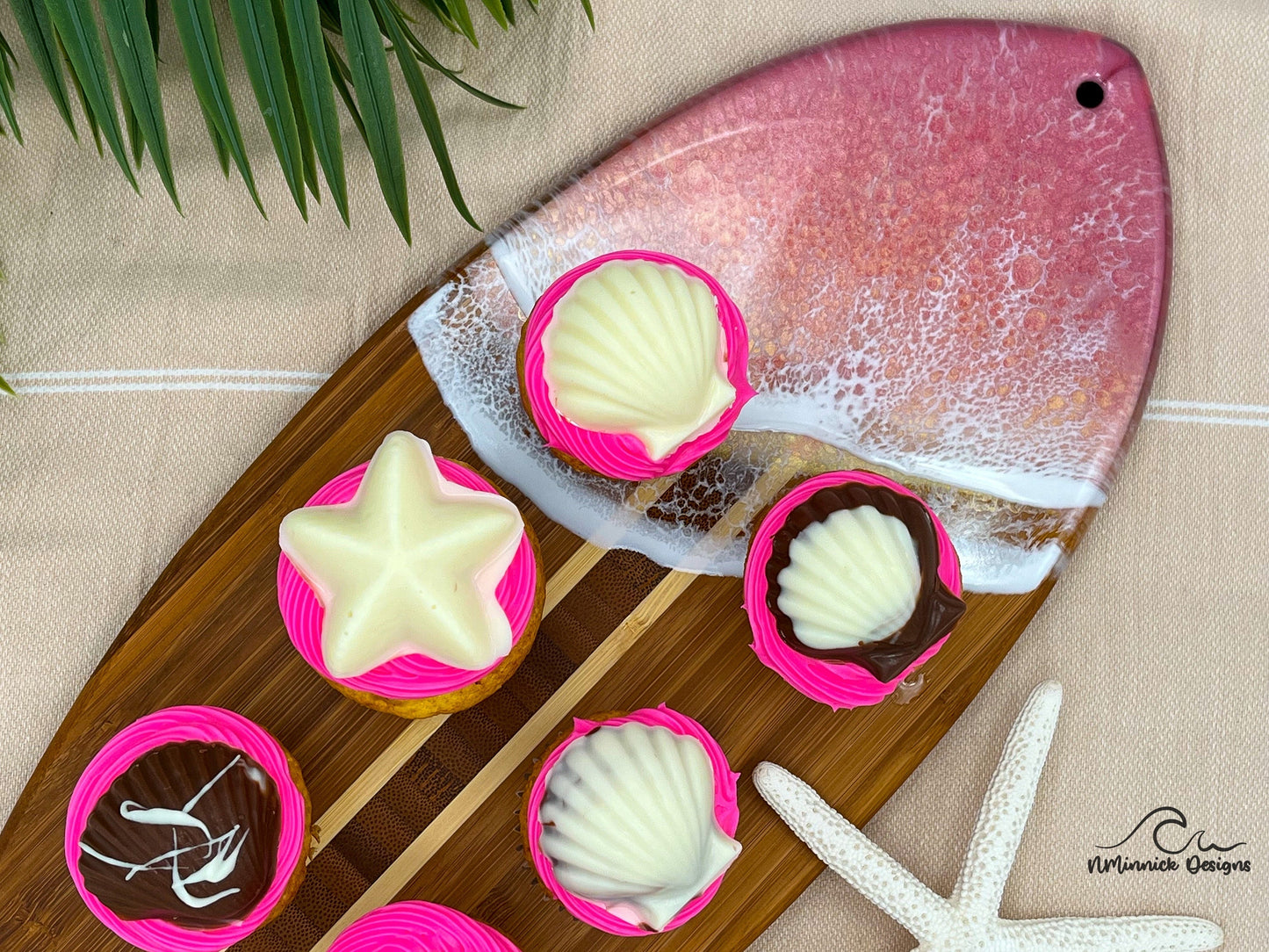 Surfboard-shaped bamboo cutting board with the top third of the board covered in a coral pink and gold tinted ocean resin art resembling the waves of the ocean. Laying on a tan beach towel next to a natural starfish (not included). Decorated with cupcakes with pink frosting and adorned with chocolate seashells