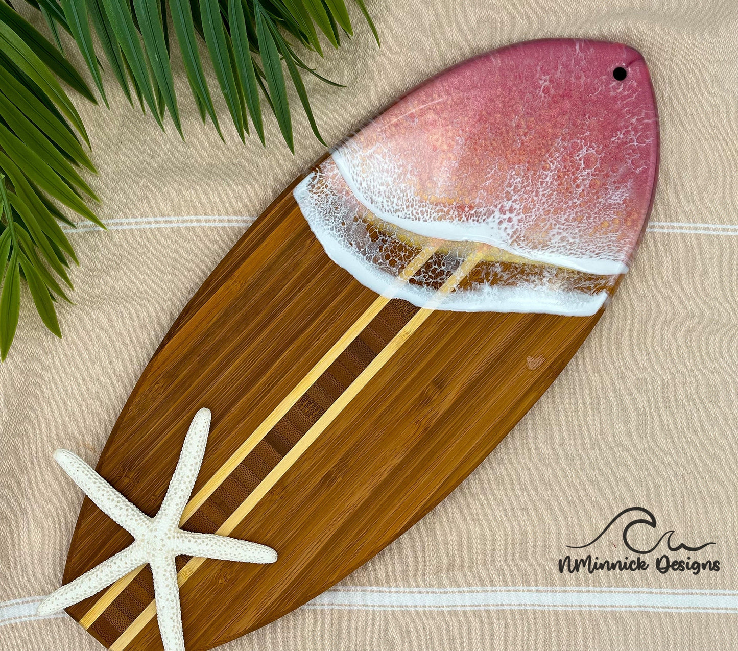 Surfboard-shaped bamboo cutting board with the top third of the board covered in a coral pink and gold tinted ocean resin art resembling the waves of the ocean.  Laying on a tan beach towel next to a natural starfish (not included).